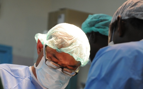 Two medical professionals are performing a surgical procedure in an operating room setting. The primary surgeon, wearing glasses, a surgical cap, face mask, and blue gown, is actively operating with surgical instruments. A second person in blue surgical attire assists from the right side.