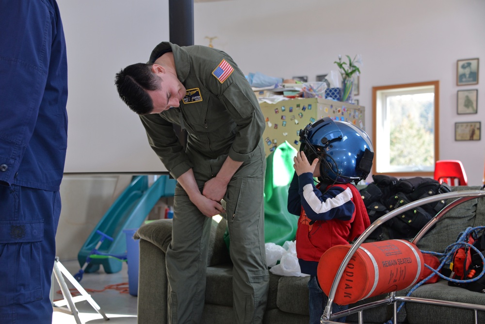 A military aviator in an olive green flight suit with a US flag patch is bending down to interact with a young child who is trying on a large blue flight helmet. The scene appears to take place in a residential or community space with children's toys visible in the background. Aviation equipment including what appears to be a red cylindrical object labeled 'KODIAK' with 'REMAIN SEATED' text is visible in the foreground.