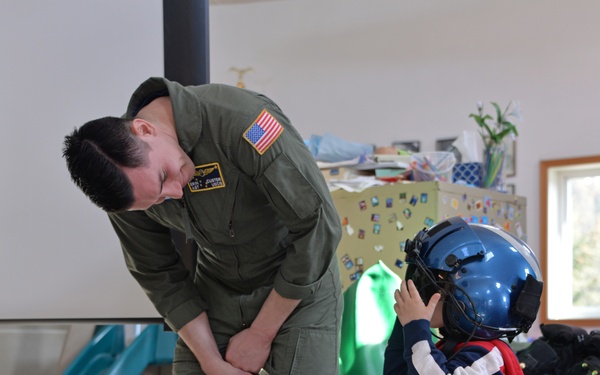 A military aviator in an olive green flight suit with a US flag patch is bending down to interact with a young child who is trying on a large blue flight helmet. The scene appears to take place in a residential or community space with children's toys visible in the background. Aviation equipment including what appears to be a red cylindrical object labeled 'KODIAK' with 'REMAIN SEATED' text is visible in the foreground.