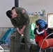 A military aviator in an olive green flight suit with a US flag patch is bending down to interact with a young child who is trying on a large blue flight helmet. The scene appears to take place in a residential or community space with children's toys visible in the background. Aviation equipment including what appears to be a red cylindrical object labeled 'KODIAK' with 'REMAIN SEATED' text is visible in the foreground.