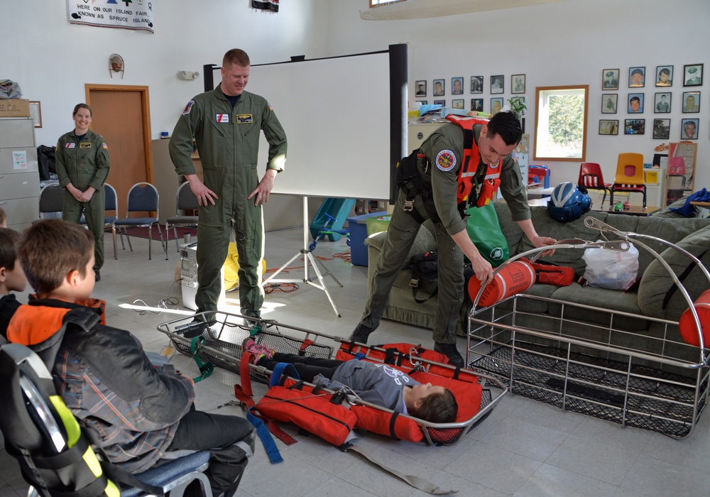 Military or Coast Guard personnel in olive green flight suits are conducting an educational demonstration for children in what appears to be a community hall or school room. One person is lying in a rescue litter/Stokes basket on the floor while another crew member demonstrates rescue equipment. A banner visible in the background references 'Spruce Island,' suggesting a remote community outreach event.