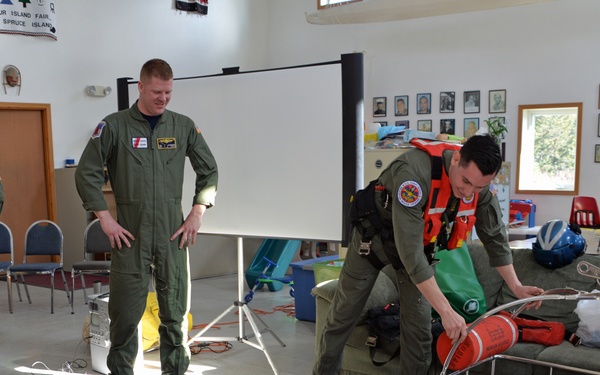 Military or Coast Guard personnel in olive green flight suits are conducting an educational demonstration for children in what appears to be a community hall or school room. One person is lying in a rescue litter/Stokes basket on the floor while another crew member demonstrates rescue equipment. A banner visible in the background references 'Spruce Island,' suggesting a remote community outreach event.