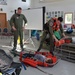 Military or Coast Guard personnel in olive green flight suits are conducting an educational demonstration for children in what appears to be a community hall or school room. One person is lying in a rescue litter/Stokes basket on the floor while another crew member demonstrates rescue equipment. A banner visible in the background references 'Spruce Island,' suggesting a remote community outreach event.