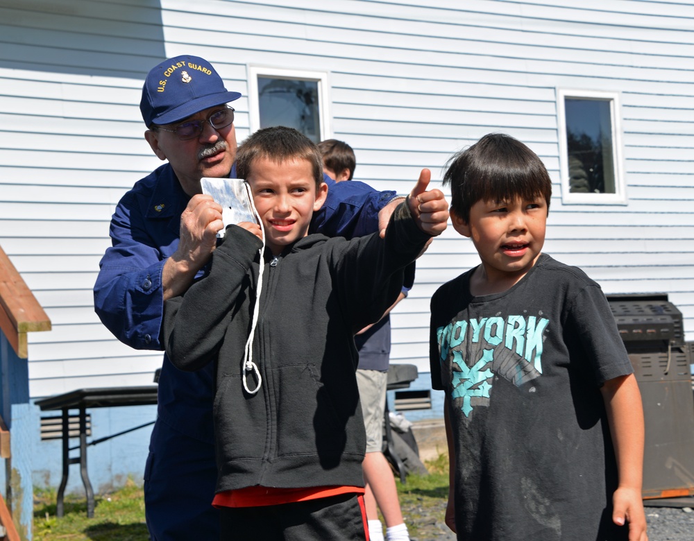 A U.S. Coast Guard member in uniform interacts with two young boys outdoors near a light blue building. One boy is giving a thumbs-up gesture while the Coast Guard member holds what appears to be a small card or photo. The scene appears to be a community outreach or educational event.