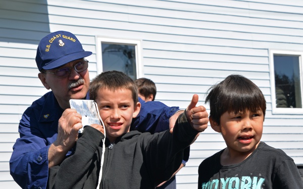 A U.S. Coast Guard member in uniform interacts with two young boys outdoors near a light blue building. One boy is giving a thumbs-up gesture while the Coast Guard member holds what appears to be a small card or photo. The scene appears to be a community outreach or educational event.