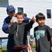 A U.S. Coast Guard member in uniform interacts with two young boys outdoors near a light blue building. One boy is giving a thumbs-up gesture while the Coast Guard member holds what appears to be a small card or photo. The scene appears to be a community outreach or educational event.