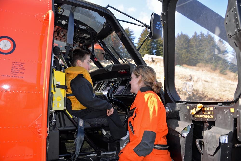 A child wearing a yellow life vest sits in the cockpit of an orange search-and-rescue helicopter while an adult in an orange flight suit speaks with them. The setting appears to be an outdoor public event or demonstration, likely allowing civilians to explore the aircraft. The helicopter's interior instruments and safety markings are clearly visible.