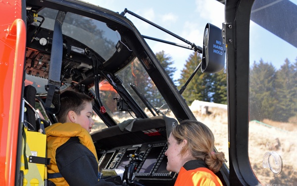 A child wearing a yellow life vest sits in the cockpit of an orange search-and-rescue helicopter while an adult in an orange flight suit speaks with them. The setting appears to be an outdoor public event or demonstration, likely allowing civilians to explore the aircraft. The helicopter's interior instruments and safety markings are clearly visible.