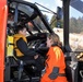 A child wearing a yellow life vest sits in the cockpit of an orange search-and-rescue helicopter while an adult in an orange flight suit speaks with them. The setting appears to be an outdoor public event or demonstration, likely allowing civilians to explore the aircraft. The helicopter's interior instruments and safety markings are clearly visible.