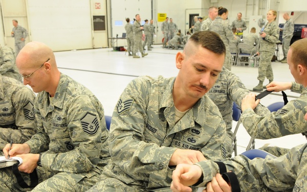 A group of U.S. Air Force personnel in ABU (Airman Battle Uniform) camouflage are seated in folding chairs inside a large hangar or training facility. The foreground shows airmen appearing to practice applying tourniquets or straps to each other's arms, suggesting a first aid or combat casualty care training exercise. Additional personnel are visible standing and moving in the background of the facility.