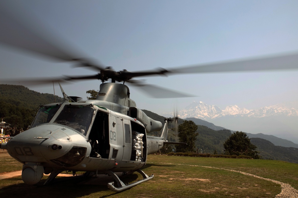 Two military helicopters are on a grassy hilltop landing zone with snow-capped mountains visible in the background. The foreground helicopter is a UH-1Y Venom (marked '08') with rotors spinning, its side door open revealing cargo or personnel inside. A larger CH-47 Chinook helicopter is visible behind it, suggesting a humanitarian or military logistics operation in a mountainous region.