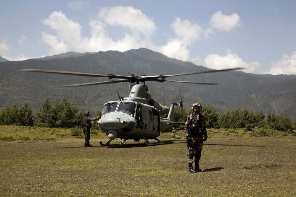 A military helicopter, appearing to be a Bell UH-1Y Venom in olive/gray livery, is landed on a grassy clearing in a mountainous, forested region. Two military personnel are visible — one standing near the helicopter's nose and another in the foreground wearing full flight gear and camouflage. The setting suggests a forward operating location or humanitarian/tactical mission in a mountainous terrain.