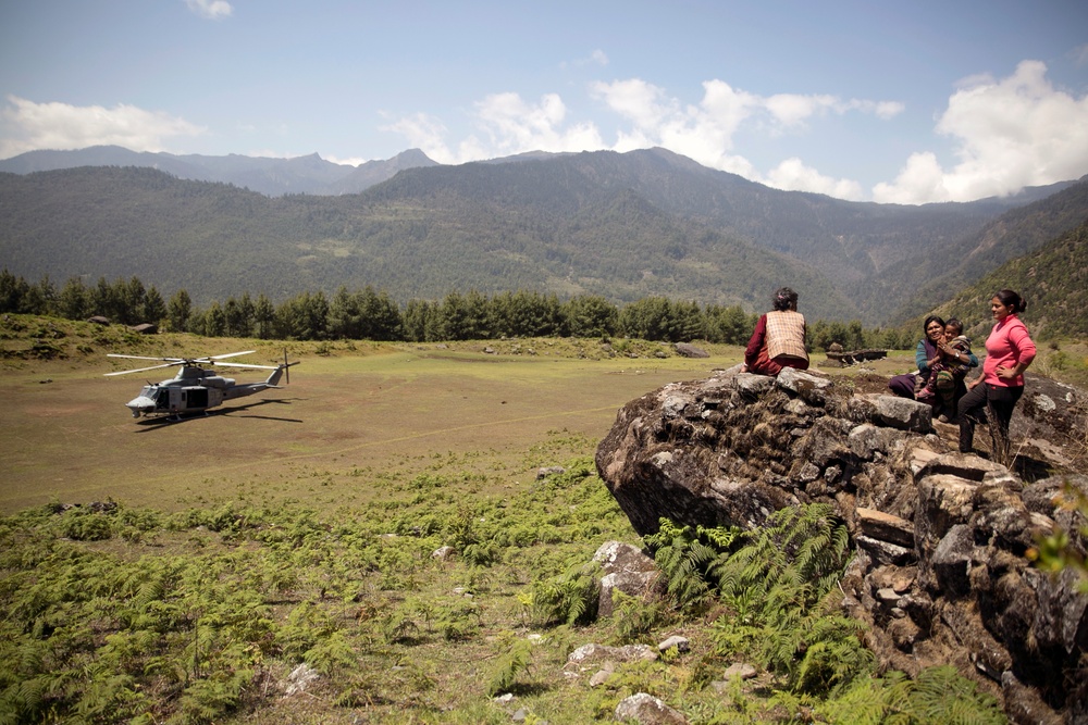 A military helicopter is landed or hovering low in a mountain valley clearing, while a small group of local civilians — including at least one woman holding an infant — observe from atop a rocky outcrop in the foreground. The setting appears to be a remote mountainous region with dense forested slopes and a broad open meadow serving as a makeshift landing zone. The scene suggests a humanitarian aid or disaster relief operation in a rural, high-altitude area.