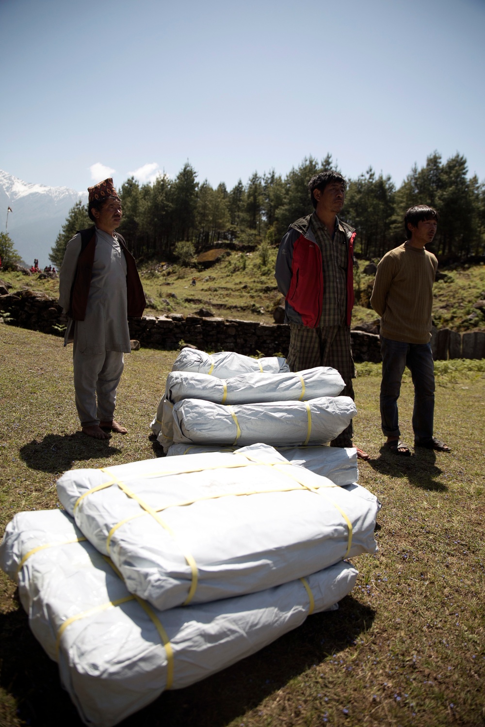Three men stand on a grassy hillside next to several large white bundles secured with yellow straps. The setting appears to be a mountainous region with pine trees and snow-capped peaks visible in the background. The scene suggests a relief supply distribution or cargo delivery operation.