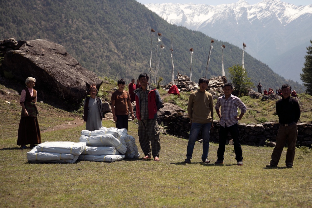 A group of approximately seven people stand on a grassy mountain meadow alongside a stack of white bundled sacks or bags. The setting appears to be a high-altitude Himalayan or similar mountain region, evidenced by snow-capped peaks in the background, forested slopes, and tall prayer flag poles. The scene suggests a supply distribution or aid delivery event in a remote mountain community.