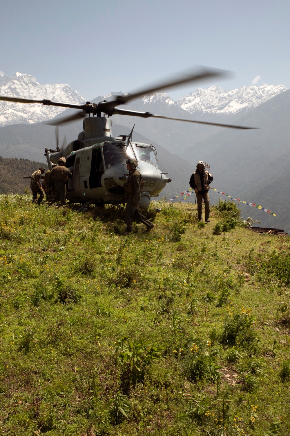 A military helicopter, identifiable as a UH-1Y Venom (tail number partially visible as '18'), has landed on a grassy highland clearing surrounded by snow-capped mountains consistent with the Himalayan range. Several personnel in military uniforms and civilian attire are gathered around the aircraft, with colorful prayer flags strung nearby suggesting a South Asian, likely Nepali, location. The scene appears to depict a humanitarian or disaster relief operation.