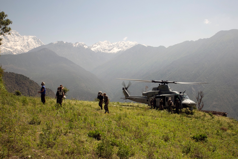A military helicopter, appearing to be a UH-1Y Venom or similar utility helicopter in gray/olive paint, has landed on a grassy mountain ridge. Several personnel in military uniforms and at least one civilian are present near the aircraft, suggesting a humanitarian relief or search-and-rescue operation. The backdrop features dramatic snow-capped mountain peaks consistent with the Himalayan range, indicating a high-altitude operational environment.