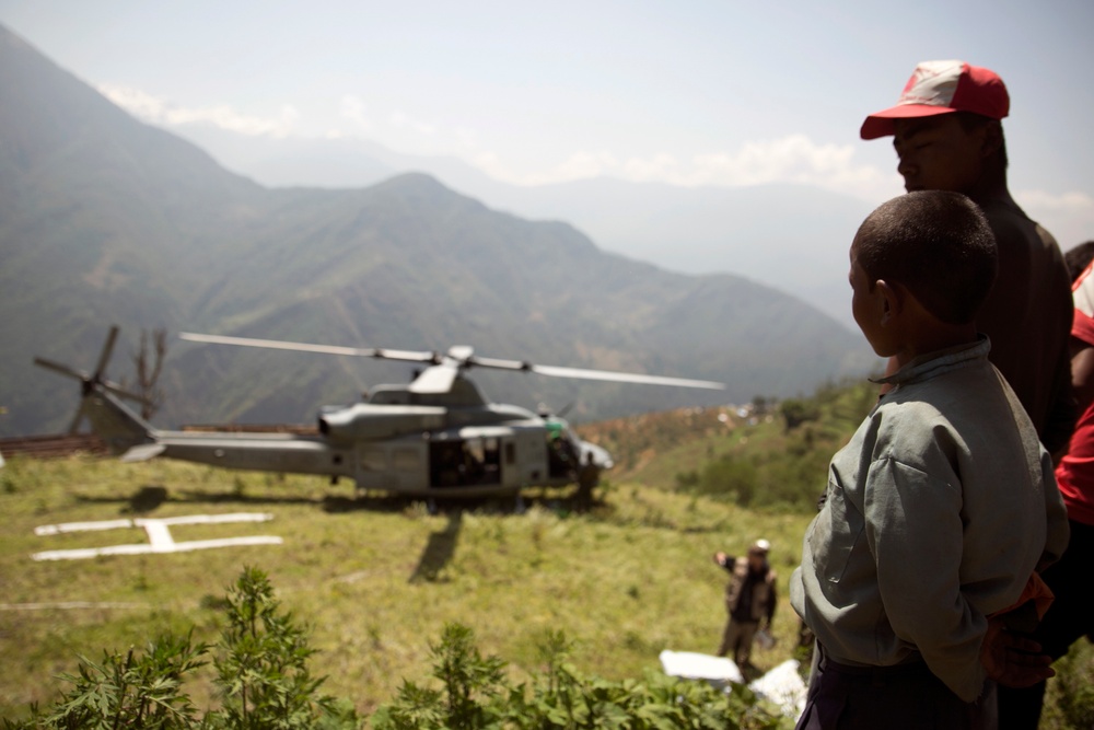 A military or humanitarian helicopter has landed on a marked landing zone in a mountainous, rural area. Local civilians, including a child and an adult wearing a red cap, observe the helicopter from a nearby hillside. The scene suggests a relief or aid operation in a remote mountain community.