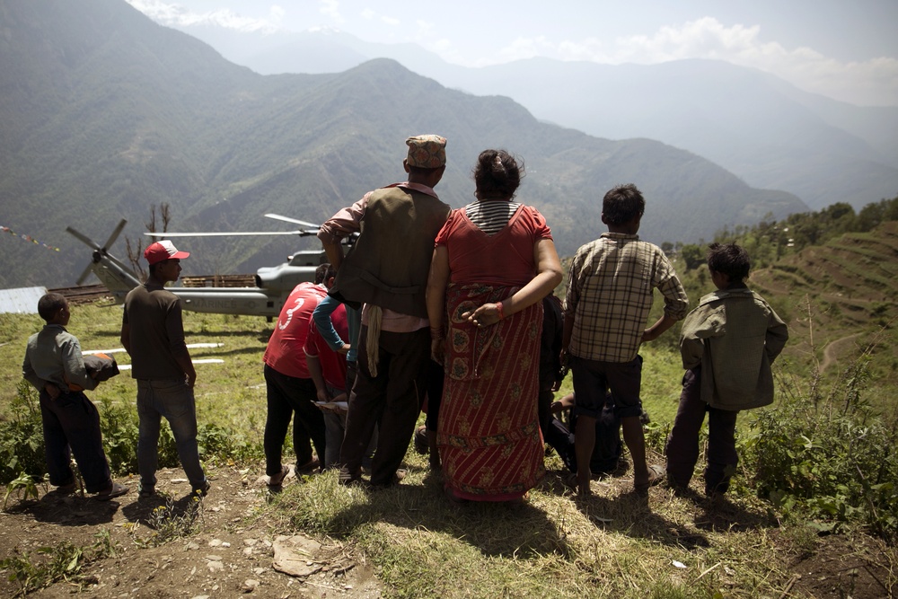 A group of people, including what appears to be a uniformed military or police officer, stand on a hillside overlooking a helicopter landed on a grassy mountain clearing. The scene appears to take place in a mountainous region consistent with the Himalayas, with terraced hillsides and deep valleys visible in the background. The context suggests a relief or evacuation operation, possibly following a natural disaster.