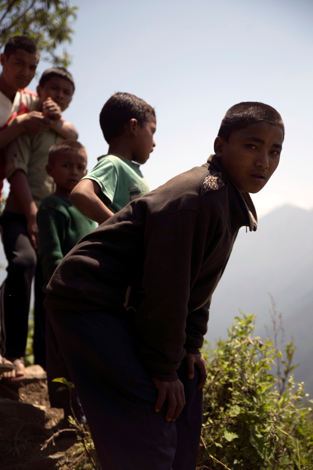 A group of children and at least one adult are gathered on what appears to be a hillside or mountain trail with a valley and mountain peaks visible in the background. The foreground features a teenage boy in a dark sweatshirt leaning forward and looking back toward the camera. The setting suggests a rural, mountainous region, likely in South or Southeast Asia.