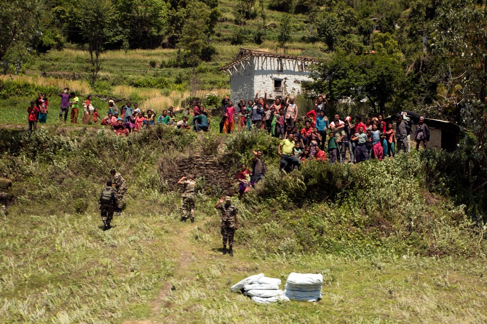 An aerial or elevated view of what appears to be a humanitarian aid distribution operation in a rural, mountainous village setting. Several uniformed personnel are walking toward a large crowd of villagers, including many children, gathered near a partially damaged stone building. White supply sacks or bags are visible on the ground in the foreground, suggesting a relief supply drop or delivery.