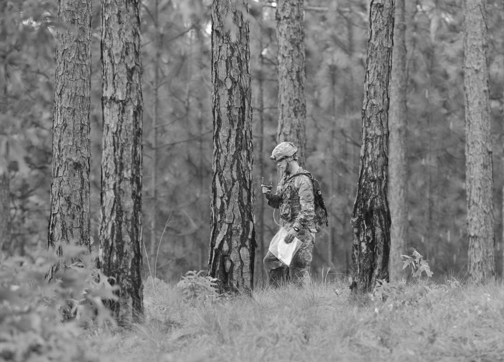 A single soldier in full combat gear navigates through a pine forest, holding what appears to be a map or document in one hand and a small device in the other. The black-and-white photograph captures the soldier mid-stride among tall, evenly spaced pine trees with low ground vegetation. The scene suggests a land navigation exercise or field training operation.