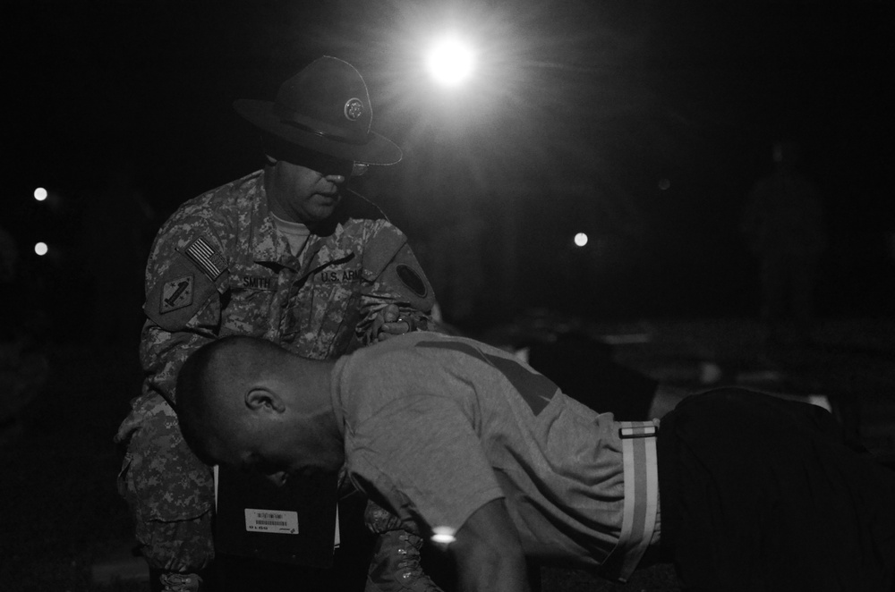 A black-and-white nighttime image showing a U.S. Army soldier in ACU (Army Combat Uniform) wearing a drill sergeant hat, appearing to supervise or assist another individual who is bent forward. The name tape 'SMITH' and 'U.S. ARMY' are visible on the uniformed soldier's chest. The scene is illuminated by a bright light source from behind, suggesting a training or field exercise environment.