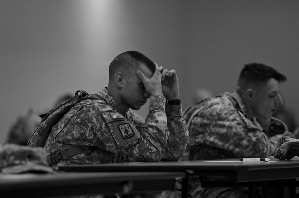 A black and white photograph showing military personnel seated at tables, likely in a classroom or briefing setting. The foreground soldier, wearing US Army ACU (Army Combat Uniform) with a US flag patch and unit patch on the left shoulder, has his head bowed with his hand raised to his forehead, suggesting concentration, stress, or fatigue. Additional soldiers are visible in the background, slightly out of focus.