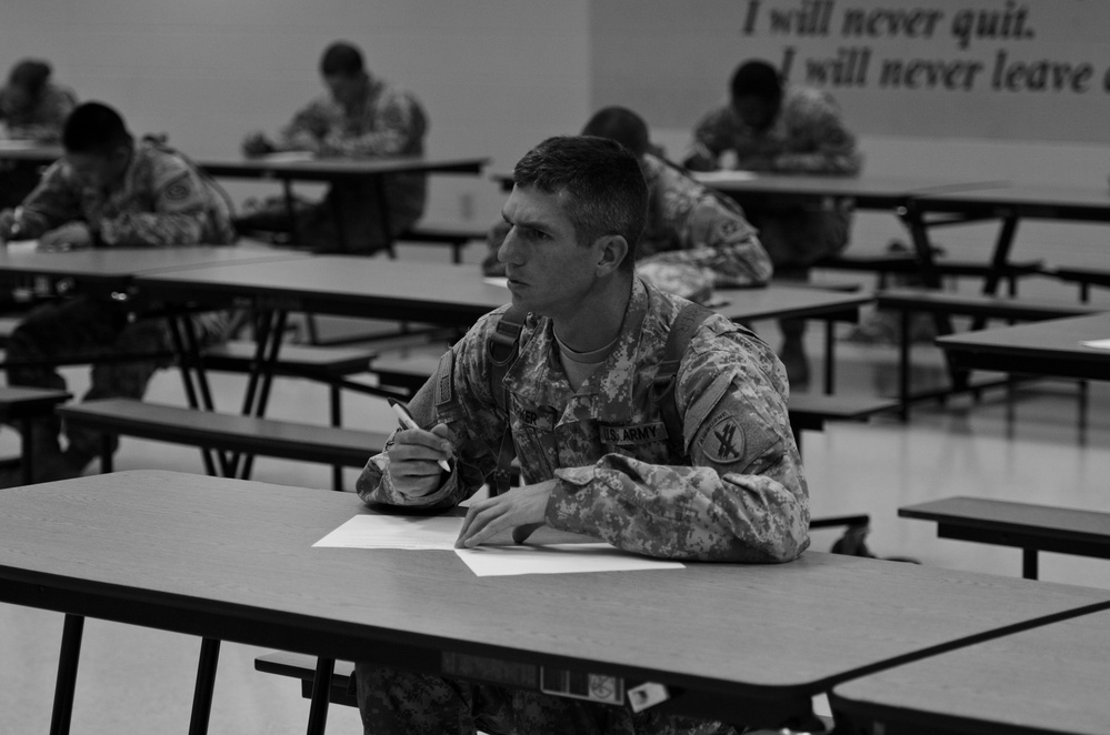 A black-and-white photograph showing U.S. Army soldiers seated at individual desks in what appears to be a classroom or testing environment. The foreground soldier, wearing ACU (Army Combat Uniform) with a U.S. Army patch, is holding a pen and looking forward attentively. A motivational phrase 'I will never quit. I will never leave' is visible on the wall in the background.
