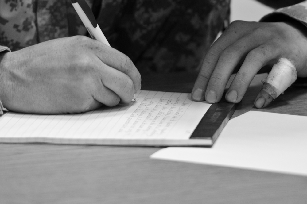 A black and white close-up photograph shows a person in military camouflage uniform writing in a lined notebook on a wooden table. The right hand holds a pen and writes on the page, while the left hand steadies the notebook using a ruler; notably, the left thumb appears to be bandaged. Additional papers are visible on the table beside the notebook.