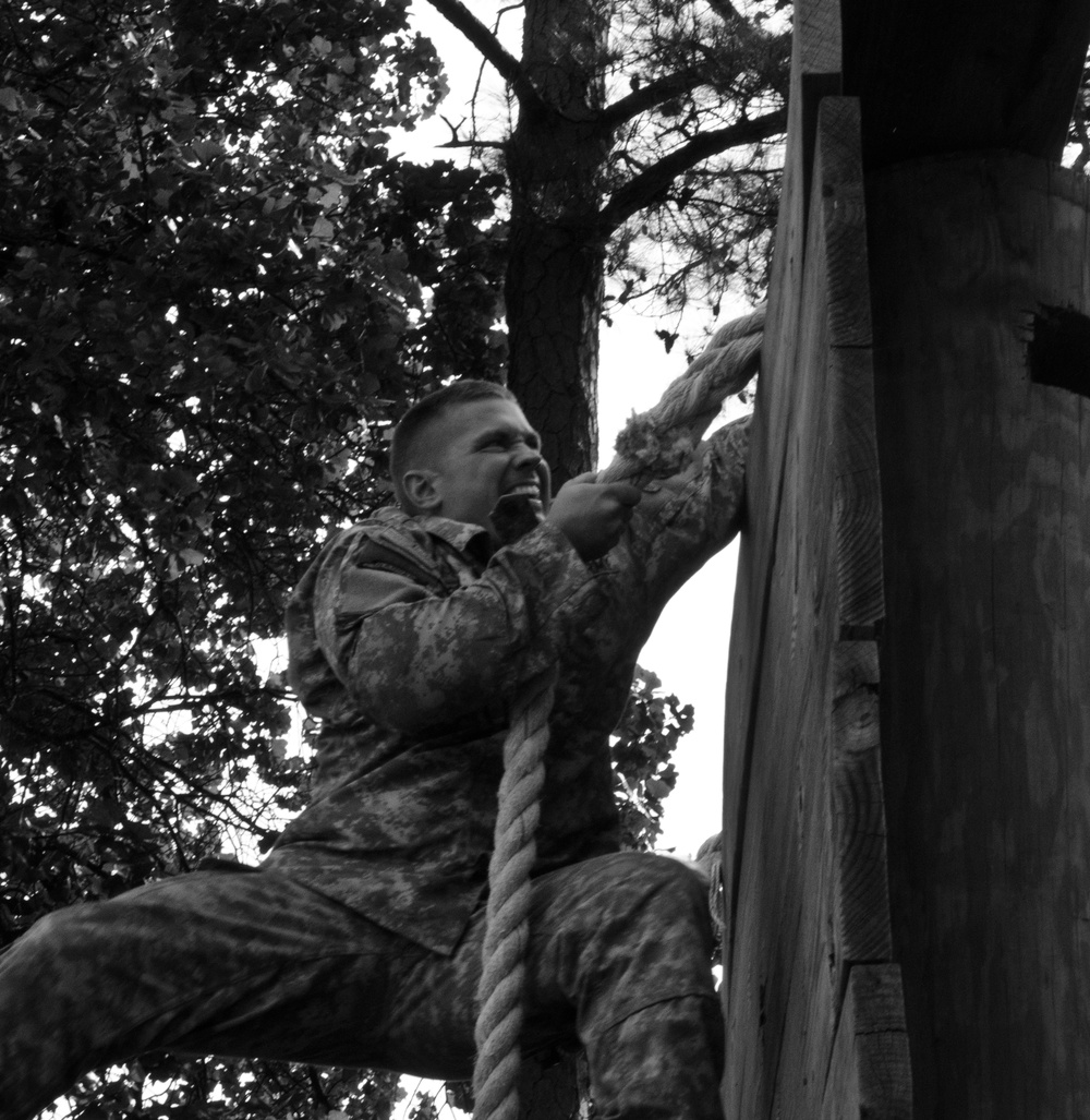 A black and white photograph showing a male soldier in camouflage uniform climbing a thick rope on what appears to be a military obstacle course. The individual is exerting visible physical effort, gripping the rope with both hands near a large wooden post or platform structure. Trees are visible in the background, suggesting an outdoor training environment.