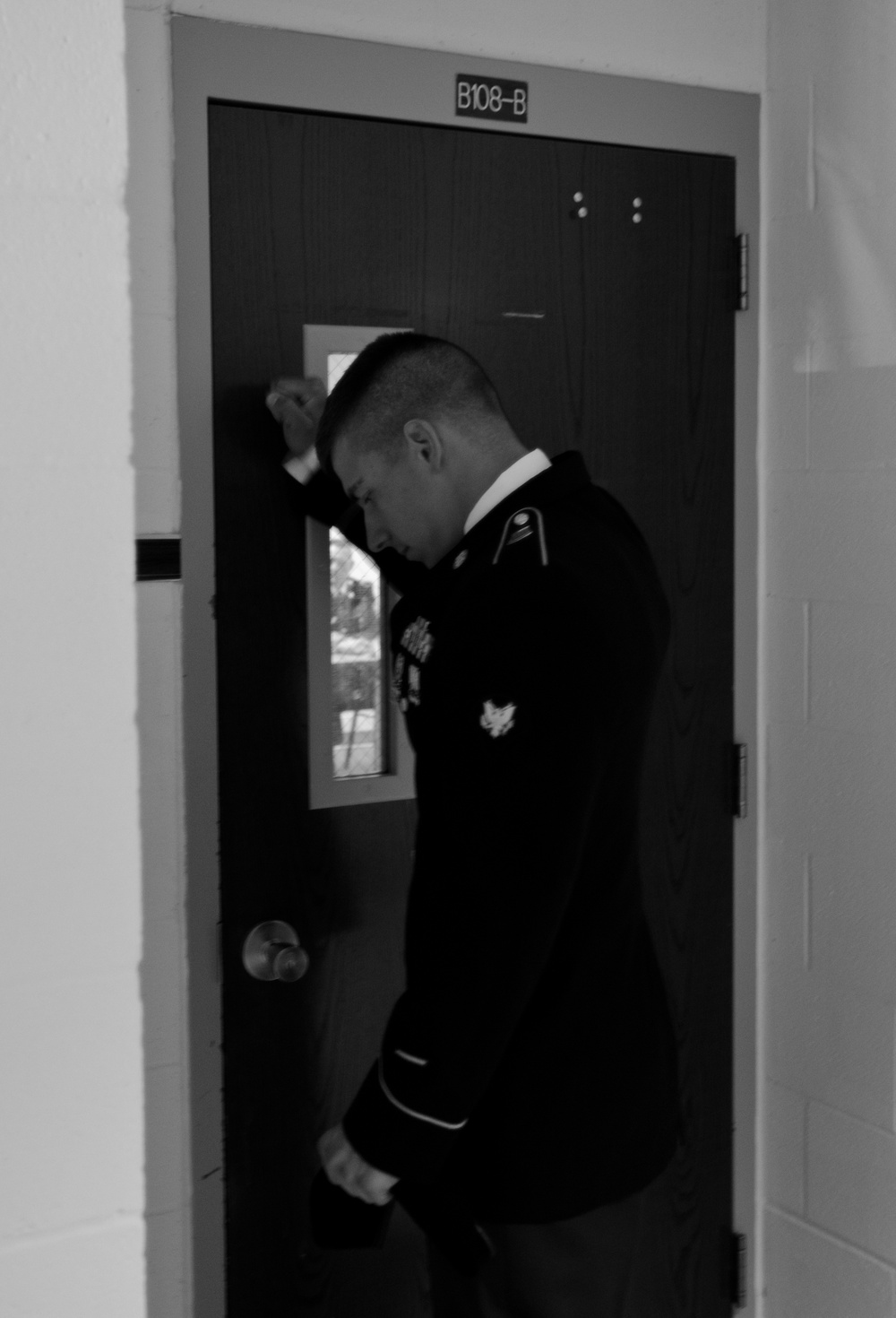 A black-and-white photograph shows a male individual wearing what appears to be a U.S. Army dress uniform, leaning with his forehead against a dark door marked 'B108-B'. His posture — head bowed and fist raised against the door — conveys a sense of distress, exhaustion, or emotional weight. The setting appears to be an institutional or military building corridor.