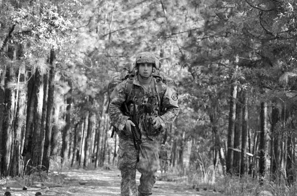 A black-and-white photograph of a single soldier in full combat gear moving along a forested trail. The soldier is wearing a combat helmet, camouflage uniform, tactical vest, and carrying a rifle while bearing a large rucksack. The setting appears to be a wooded training or operational environment with tall pine trees lining both sides of the path.