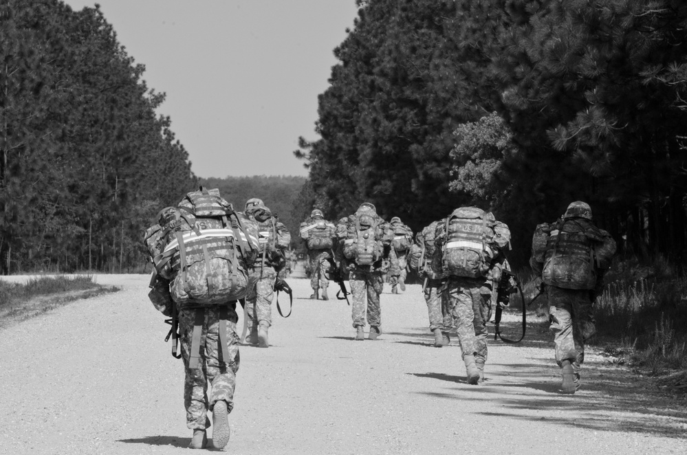 A black-and-white photograph shows a column of military personnel conducting a road march along a straight gravel or paved road flanked by tall pine trees. The soldiers are moving away from the camera, all carrying large rucksacks and wearing combat uniforms and helmets. The image appears to depict a training exercise or tactical movement, likely at a U.S. military installation.