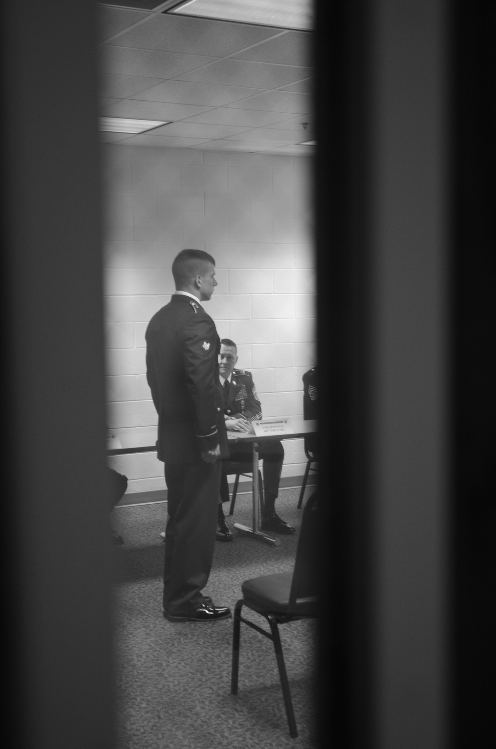 A black-and-white photograph taken through a window or door frame showing what appears to be a military board or evaluation proceeding. One uniformed soldier stands at attention while at least two others are seated at a table, suggesting a formal review or promotion board setting. The image is framed by vertical dark elements consistent with a door or window frame.