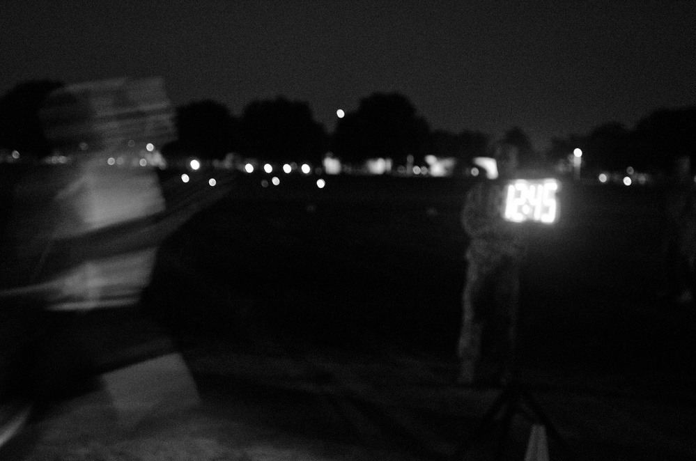 A nighttime outdoor scene captured with significant motion blur and low light conditions. The image shows silhouetted figures and an illuminated digital display or scoreboard in the background, surrounded by distant lights and tree silhouettes. The overall image quality is very low due to darkness and camera movement.