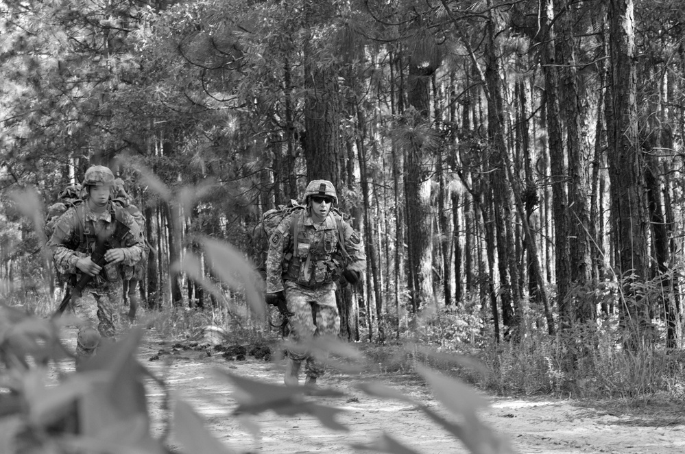 A black-and-white photograph showing two soldiers in full combat gear moving along a sandy trail through a pine forest. Both are wearing helmets, body armor, and carrying large rucksacks, suggesting a field training exercise or patrol. The foreground contains blurred foliage, giving the image a sense of depth and tactical perspective.