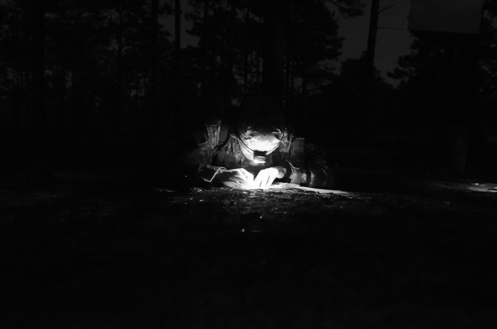 A black and white nighttime photograph showing a single individual illuminated by a headlamp or mounted light source in an outdoor wooded setting. The person appears to be crouched low to the ground, focused on something directly in front of them. The surrounding environment is nearly completely dark, with tree silhouettes faintly visible in the background.