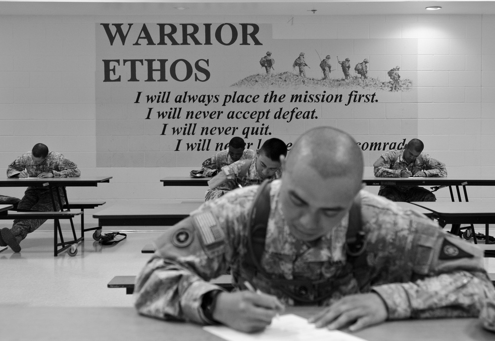 A black-and-white photograph shows multiple soldiers in camouflage uniforms seated at tables and writing, likely completing a written examination or assessment. The setting appears to be a military training facility or classroom. A large 'Warrior Ethos' mural dominates the background wall, displaying the U.S. Army's Warrior Ethos creed alongside an illustration of soldiers in combat.