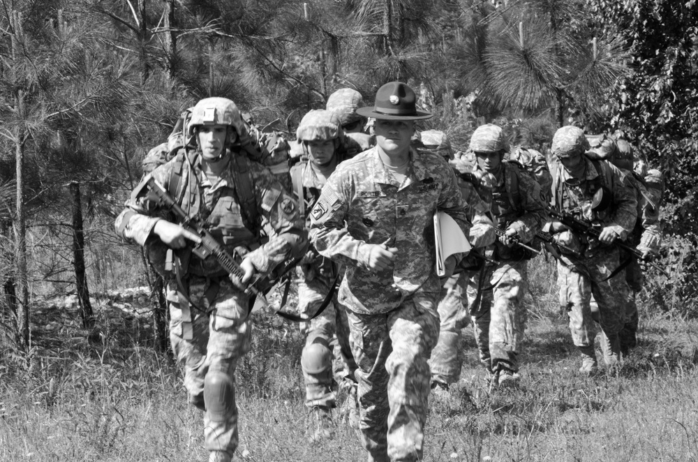 A black-and-white photograph showing a group of military personnel moving through a field at the edge of a pine forest. A central figure wearing a distinctive drill sergeant campaign cover (campaign hat) leads or accompanies soldiers carrying rifles and large rucksacks. The scene appears consistent with a military training exercise or field operation.