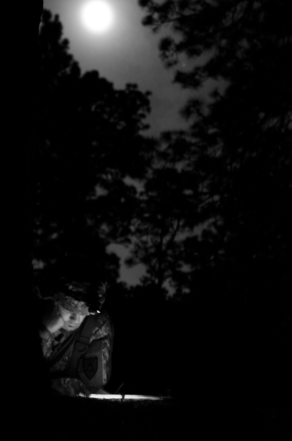 A black-and-white nighttime photograph showing a young child illuminated by a light source from below, set against a dark outdoor background with tree silhouettes and a bright full moon overhead. The child appears to be wearing camouflage or patterned clothing and is leaning over a glowing object. The contrast between the moonlight above and the artificial light below creates a dramatic compositional effect.