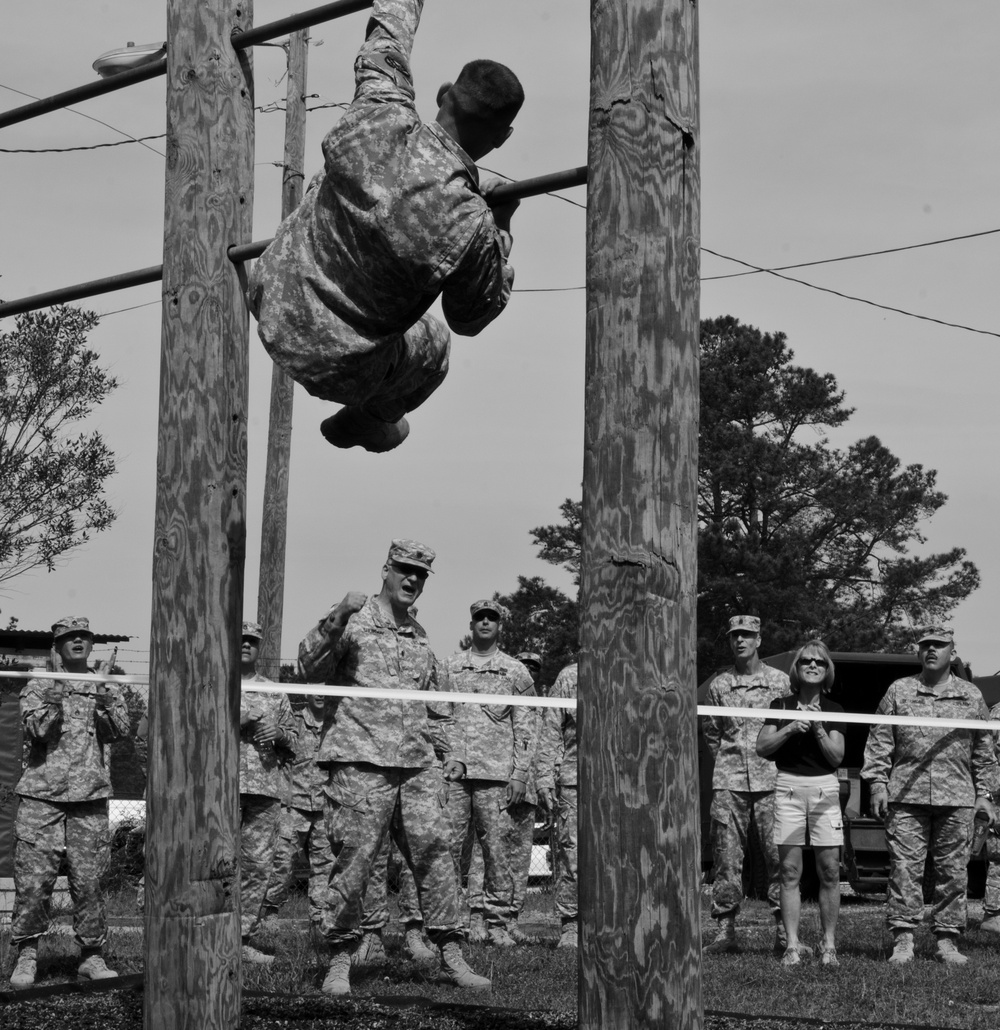 A black and white photograph showing a military soldier in camouflage uniform performing an obstacle course exercise, hanging inverted from horizontal bars between two wooden utility poles. A group of uniformed military personnel and one civilian woman observe from behind a rope barrier in the background. The setting appears to be a military training facility with trees and structures visible.
