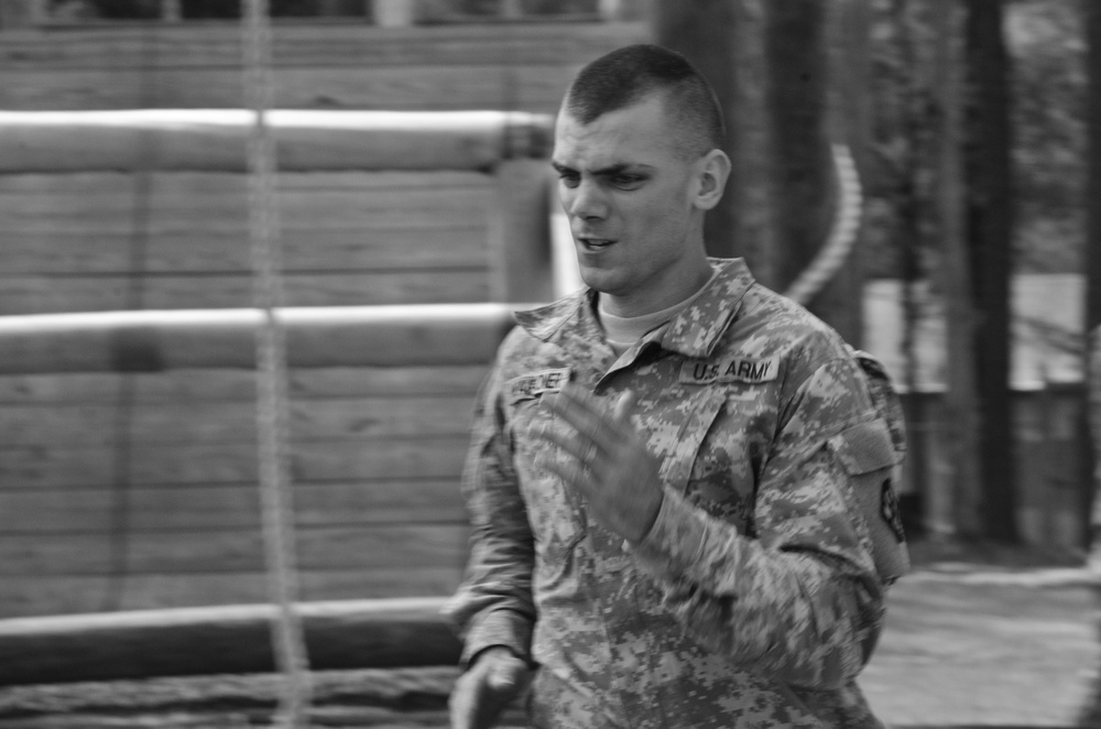 A young U.S. Army soldier in ACU (Army Combat Uniform) is captured in a black-and-white photograph, appearing to be speaking or giving instruction. He has a short military haircut and is gesturing with one hand. The background suggests a military training or base environment with wooden structures visible.