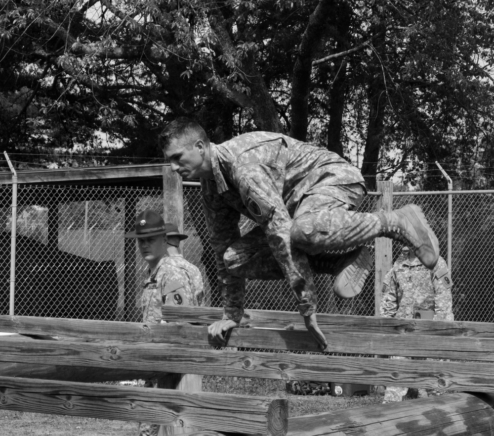 A black and white photograph showing a soldier in camouflage uniform navigating a wooden obstacle course barrier, likely during military training. Two other uniformed personnel, one wearing a distinctive drill sergeant hat, observe in the background. The setting appears to be a military installation with a chain-link fence and trees visible.
