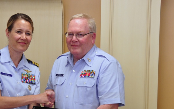 Two United States Coast Guard personnel are posing together while holding a framed Certificate of Retirement. The female officer on the left, wearing commander-rank insignia and aviator wings, appears to be presenting the certificate to the male enlisted member on the right. The ceremony appears to be taking place in an indoor facility.