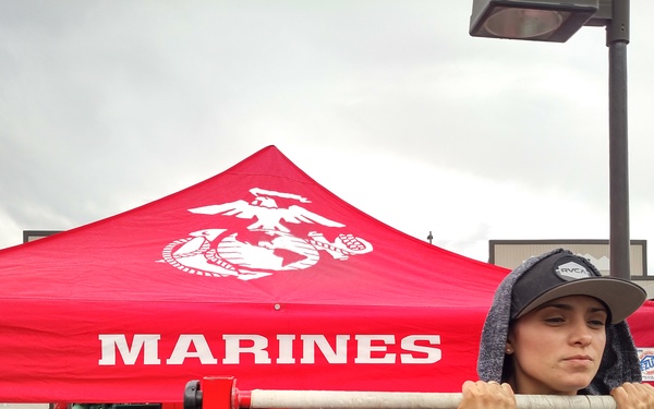 A young person wearing a RVCA snapback cap and graphic hoodie stands at a U.S. Marines recruitment booth at what appears to be an outdoor public event. They are gripping a pull-up bar mounted on a red frame, likely participating in a Marines fitness challenge. The booth features a red canopy with the USMC Eagle, Globe, and Anchor emblem, along with a branded red Hummer H2 vehicle.