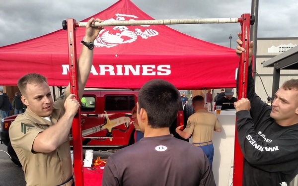 A United States Marines recruiting booth is set up at an outdoor public event, featuring a red pull-up bar challenge station under a red Marines-branded canopy tent. Two Marines — one in tan service uniform and one in a black Marines long-sleeve shirt with camouflage trousers — are assisting a civilian in dark athletic wear with a pull-up challenge. A prize board labeled 'Marine Corps Challenge' is visible listing rewards such as stickers, lanyards, hats, water bottles, and T-shirts.