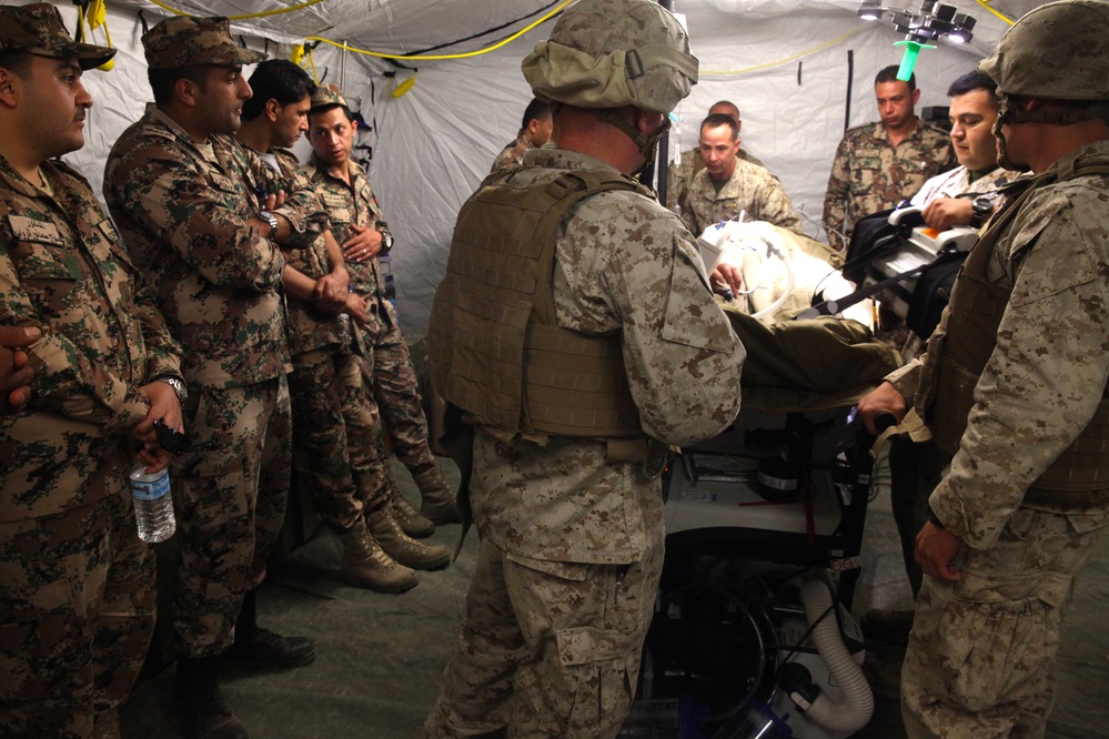 Military personnel from two distinct forces are gathered inside a field medical tent around what appears to be a medical training station or patient gurney. Soldiers in MARPAT (US Marine) digital camouflage and soldiers in a different camouflage pattern (possibly Jordanian Armed Forces) are observing a medical demonstration or procedure. The setting appears to be a joint military medical training exercise.