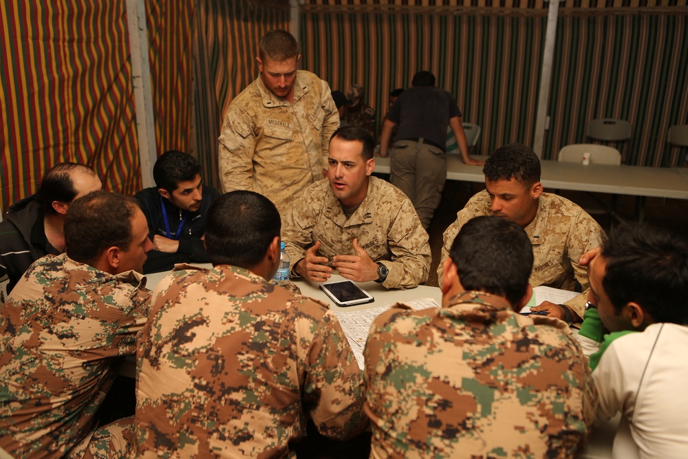 A group of military personnel from at least two different forces are gathered around a table in what appears to be a briefing or planning session inside a tent or temporary structure. U.S. Marines in desert MARPAT uniforms are engaging with personnel wearing a different digital camouflage pattern, suggesting a multinational or coalition meeting. A tablet device and documents are visible on the table, indicating a working session or training exercise.