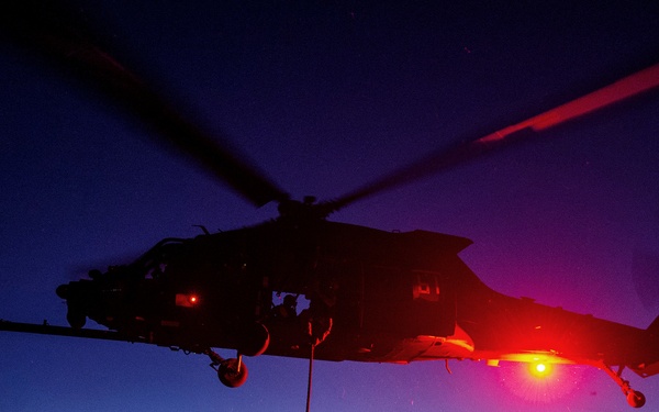 A dramatic twilight/dusk military training operation showing personnel being extracted or inserted via rope from a hovering helicopter over desert terrain. A second helicopter is visible in the background performing a similar operation. The scene is illuminated by intense red lighting from the primary helicopter, casting a vivid glow over the desert landscape.